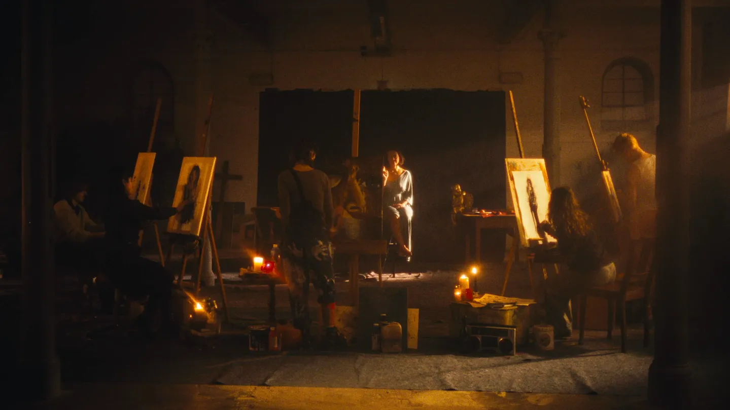 Stigma Still image: Five artists in front of their easels in a room lit only by candlelight. A few rays of sunlight fall through a small window at the top right, directly onto an elderly woman sitting in the middle as their model.